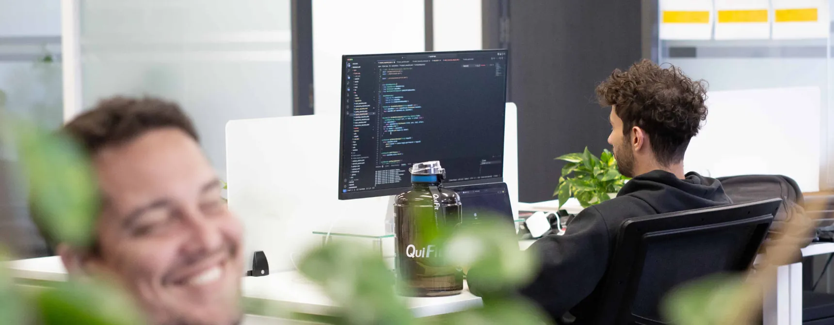 A developer at a workstation with code on screen while a colleague talks nearby